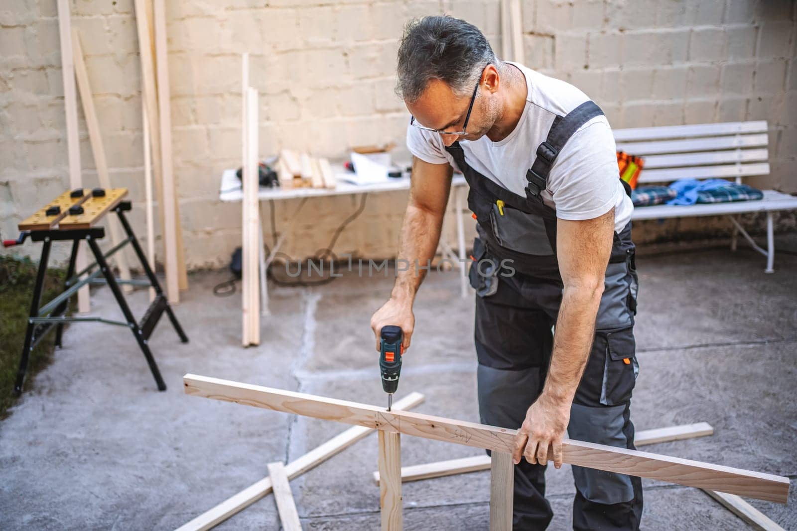 Mature male carpenter using electric screwdriver on a wooden plank outdoors.