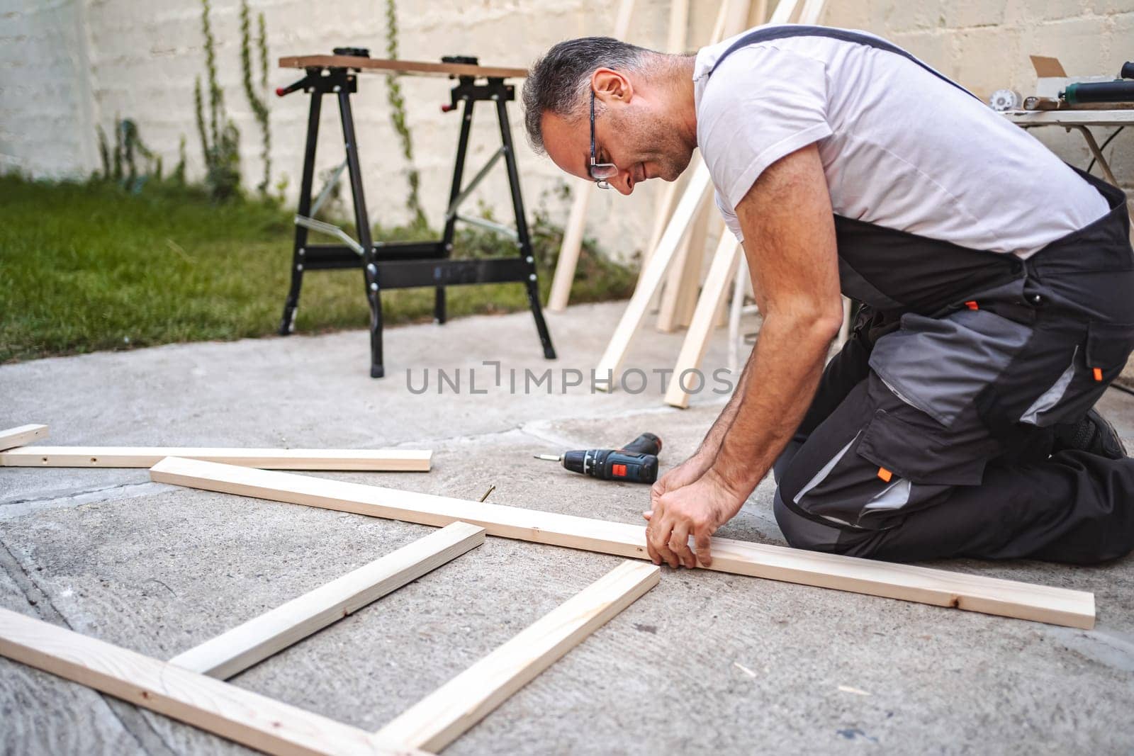 Mature male carpenter using electric screwdriver on a wooden plank outdoors.