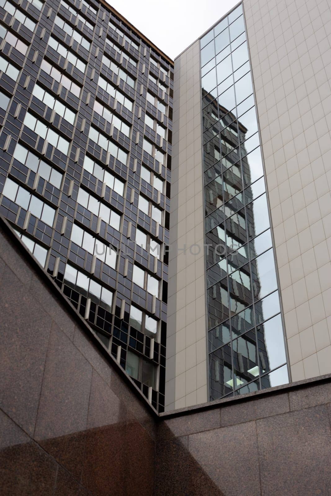 Vertical photography of corners of two high rise buildings during the exit from the underground passage. Tall office buildings facade on gray cloudy day