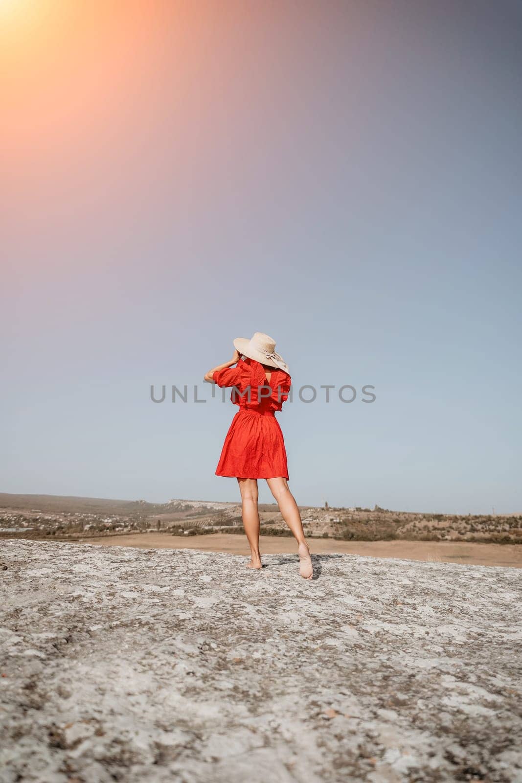 A young woman stands on a rocky outcrop wearing a red dress and a straw hat, looking out at the horizon.