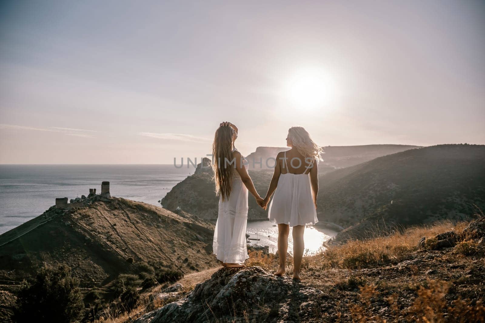 Two women are standing on a hill overlooking the ocean. They are holding hands and looking out at the water. The scene is peaceful and serene, with the sun shining brightly in the background