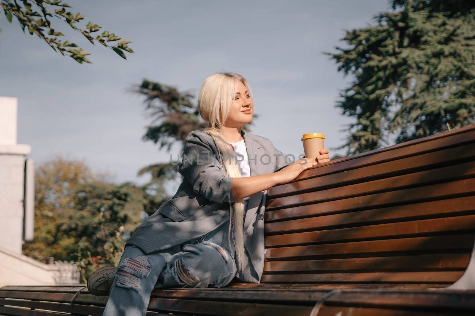 A blonde woman sits on a bench with a cup of coffee in her hand. She is wearing a gray jacket and jeans. The bench is wooden and has a curved back. The scene is peaceful and relaxing