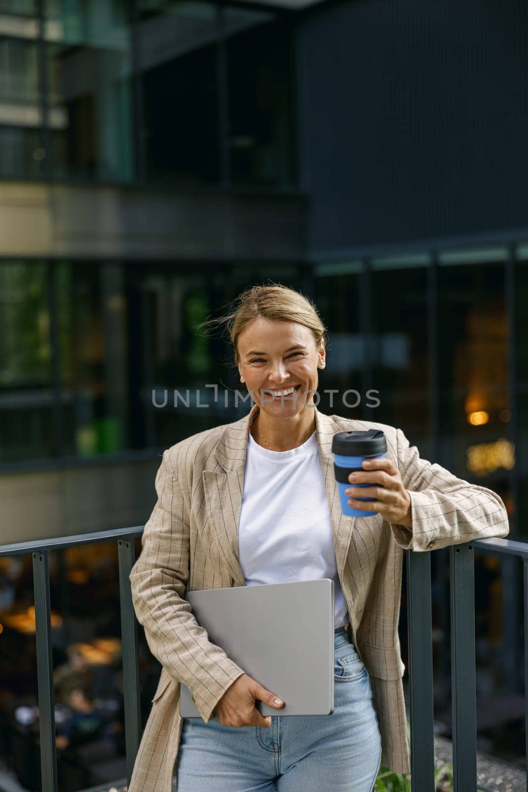 Sales manager standing with laptop and coffee on office terrace during break time and looks camera by Yaroslav_astakhov
