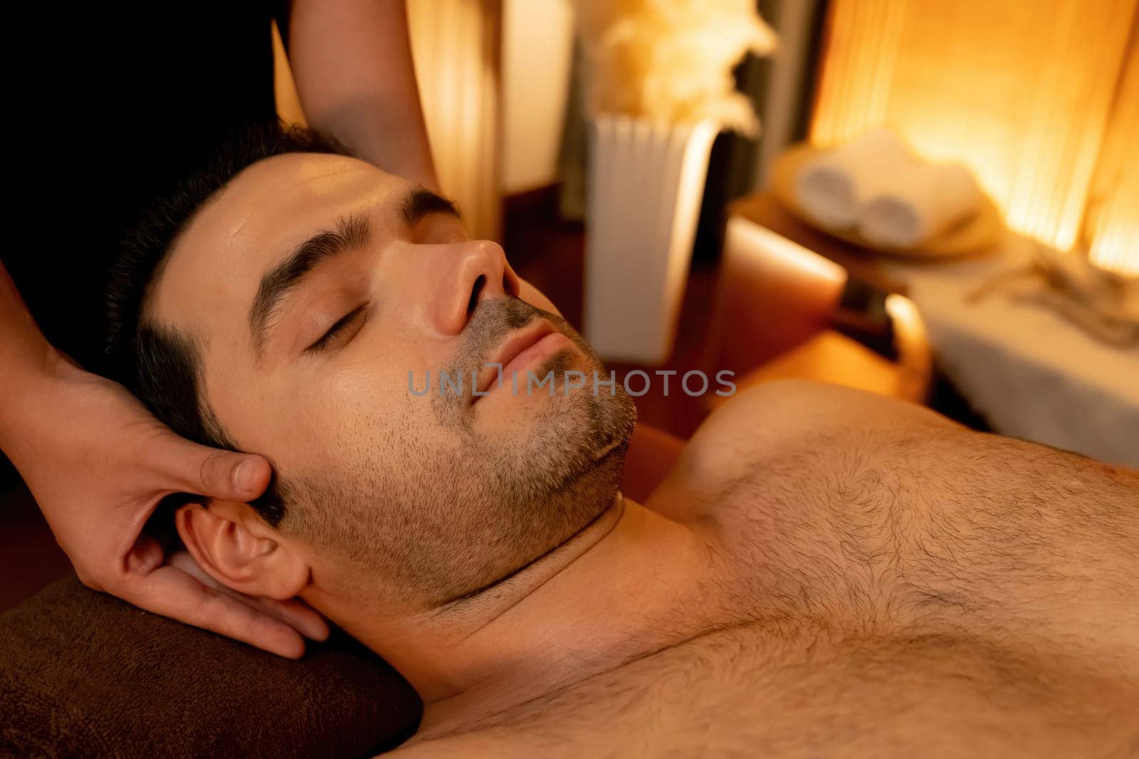 Caucasian man enjoying relaxing anti-stress head massage and pampering facial beauty skin recreation leisure in warm candle lighting ambient salon spa in luxury resort or hotel. Quiescent