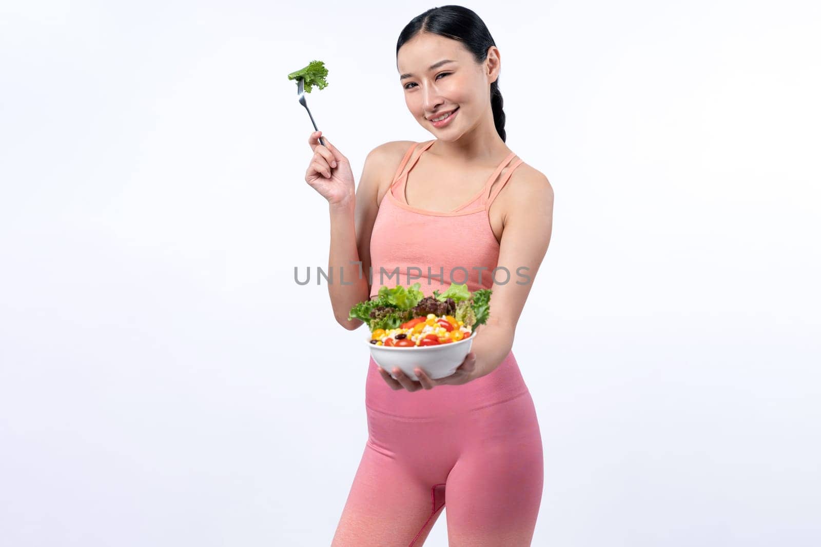 Young sporty Asian woman in sportswear holding salad bowl fill with vibrant of fruit and vegetable. Natural youthful and fit body lifestyle with balance nutrition on isolated background. Vigorous