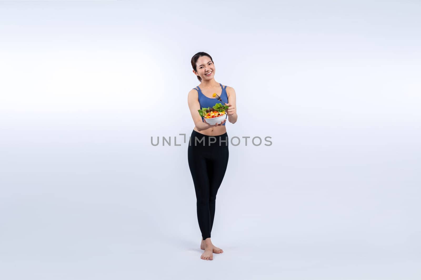 Young sporty Asian woman in sportswear holding salad bowl fill with vibrant of fruit and vegetable. Natural youthful and fit body lifestyle with balance nutrition on isolated background. Vigorous