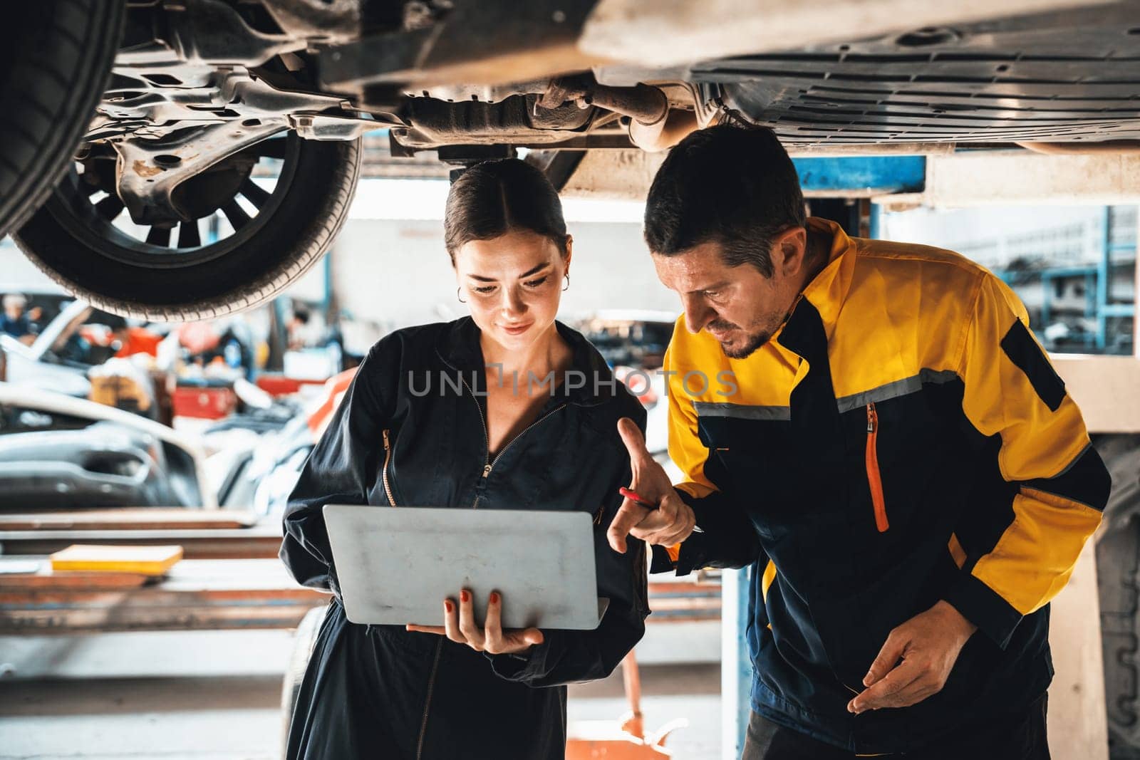 Two vehicle mechanic working together underneath lifted car, conduct car inspection with laptop. Automotive service technician in uniform carefully make diagnostic troubleshooting. Oxus