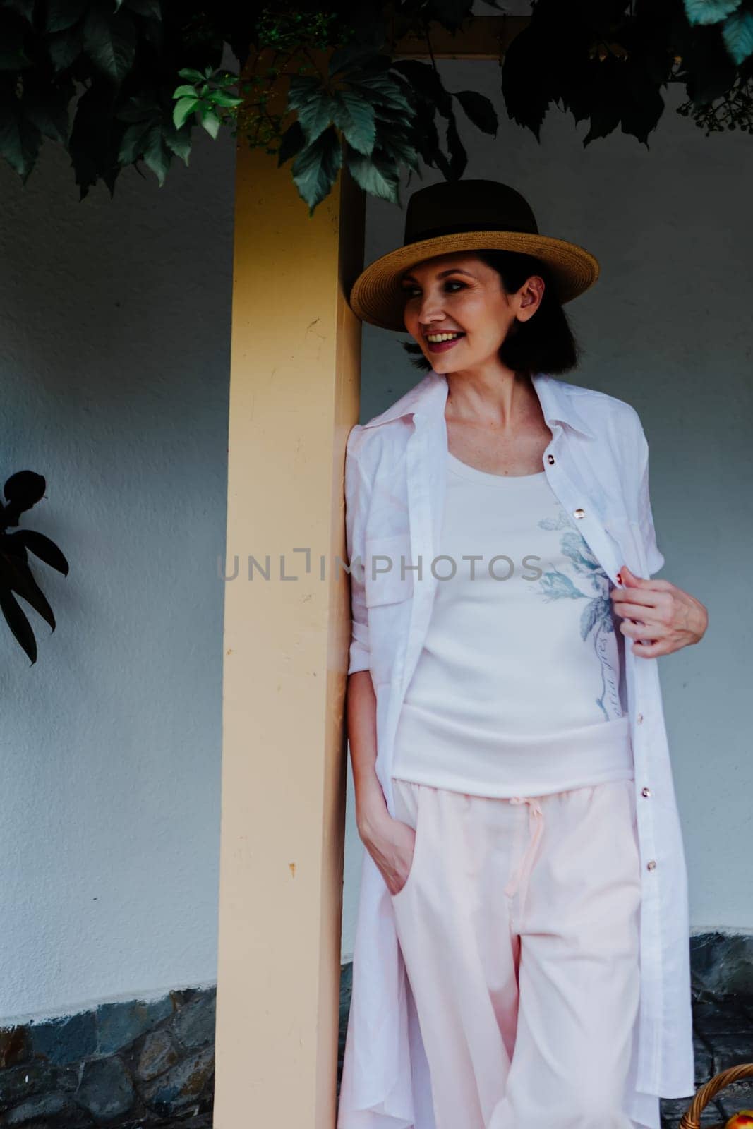 Woman dressed in casual, light-colored clothes and a wide-brimmed hat leans against a post under a leafy green canopy.