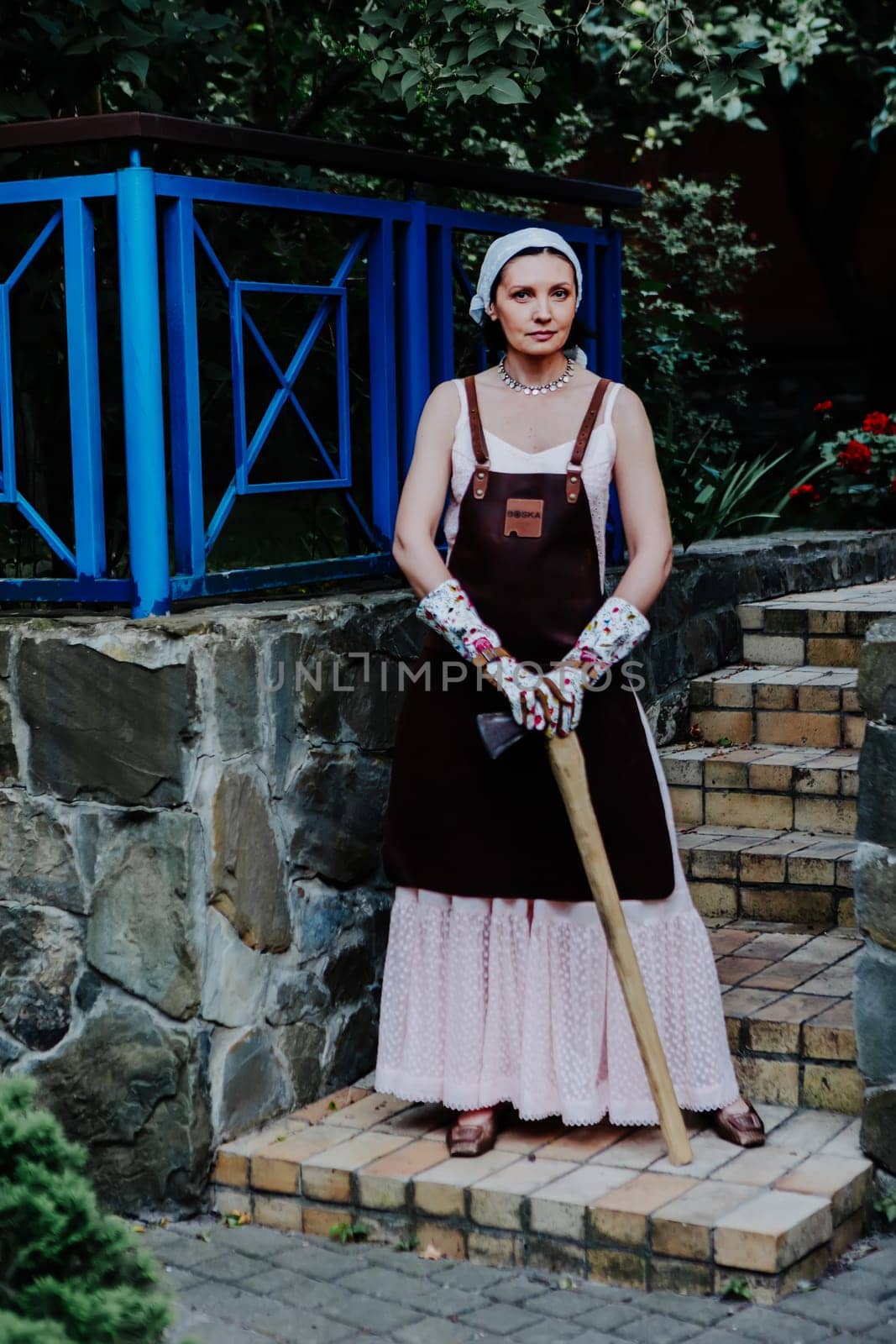 Woman dressed in a long pink dress and leather apron stands confidently against a stone wall holding axe