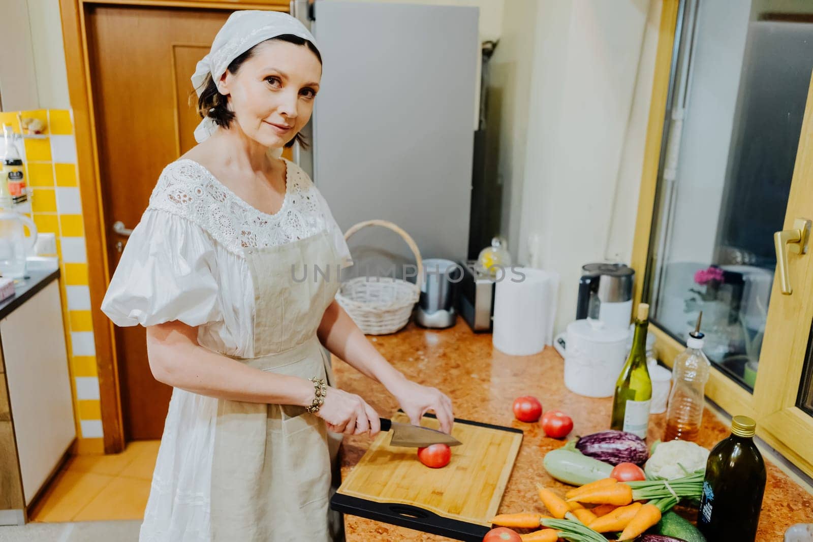 A woman is preparing a meal in a cozy kitchen. She is chopping fresh tomatoes on a wooden cutting board, surrounded by an array of colorful vegetables such as carrots, cauliflower, and cucumbers.