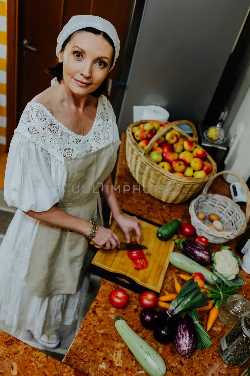 A woman is preparing a meal in a cozy kitchen. She is chopping fresh tomatoes on a wooden cutting board, surrounded by an array of colorful vegetables such as carrots, cauliflower, and cucumbers.