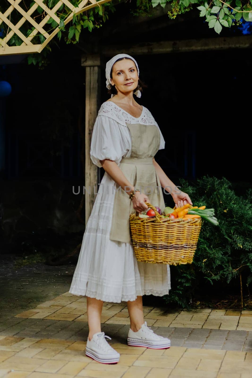 Woman dressed in a traditional white dress and apron holds a large wicker basket filled with fresh vegetables, including carrots, tomatoes, and eggplants, in a cozy kitchen.