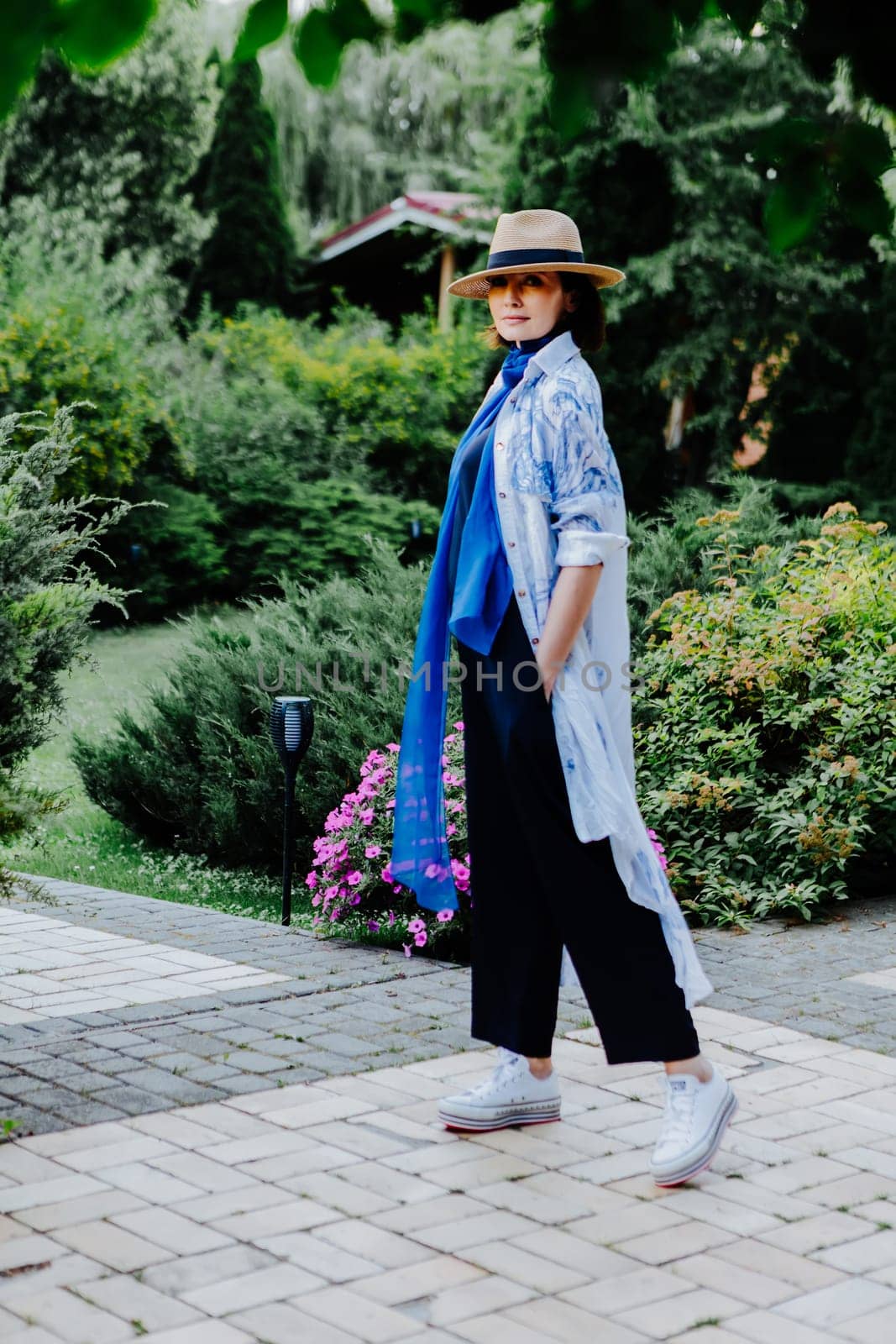 Fashionable woman is seen walking outdoors, dressed in a stylish combination of a long, flowing patterned coat, a blue scarf, wide-leg black pants, white sneakers, and a straw hat