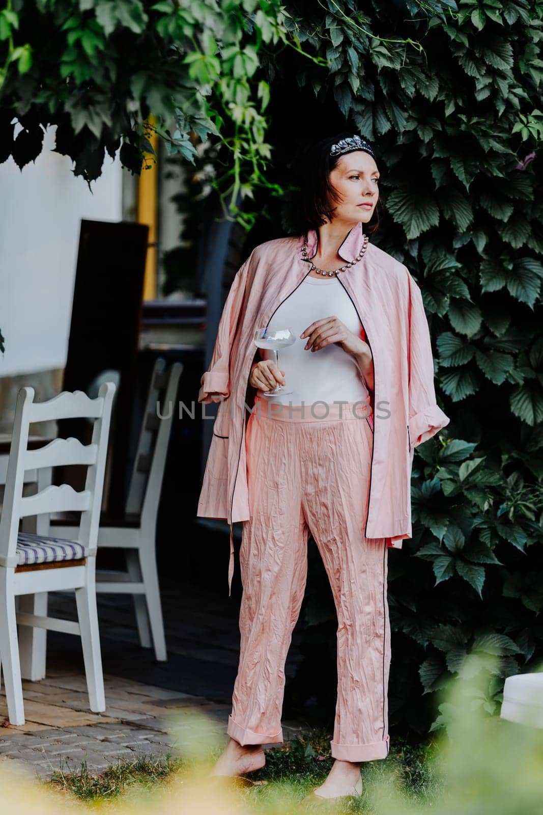Fashionable woman dressed in a stylish light pink ensemble, including a satin jacket and matching pants, is standing outdoors under a canopy of leaves, holding a glass