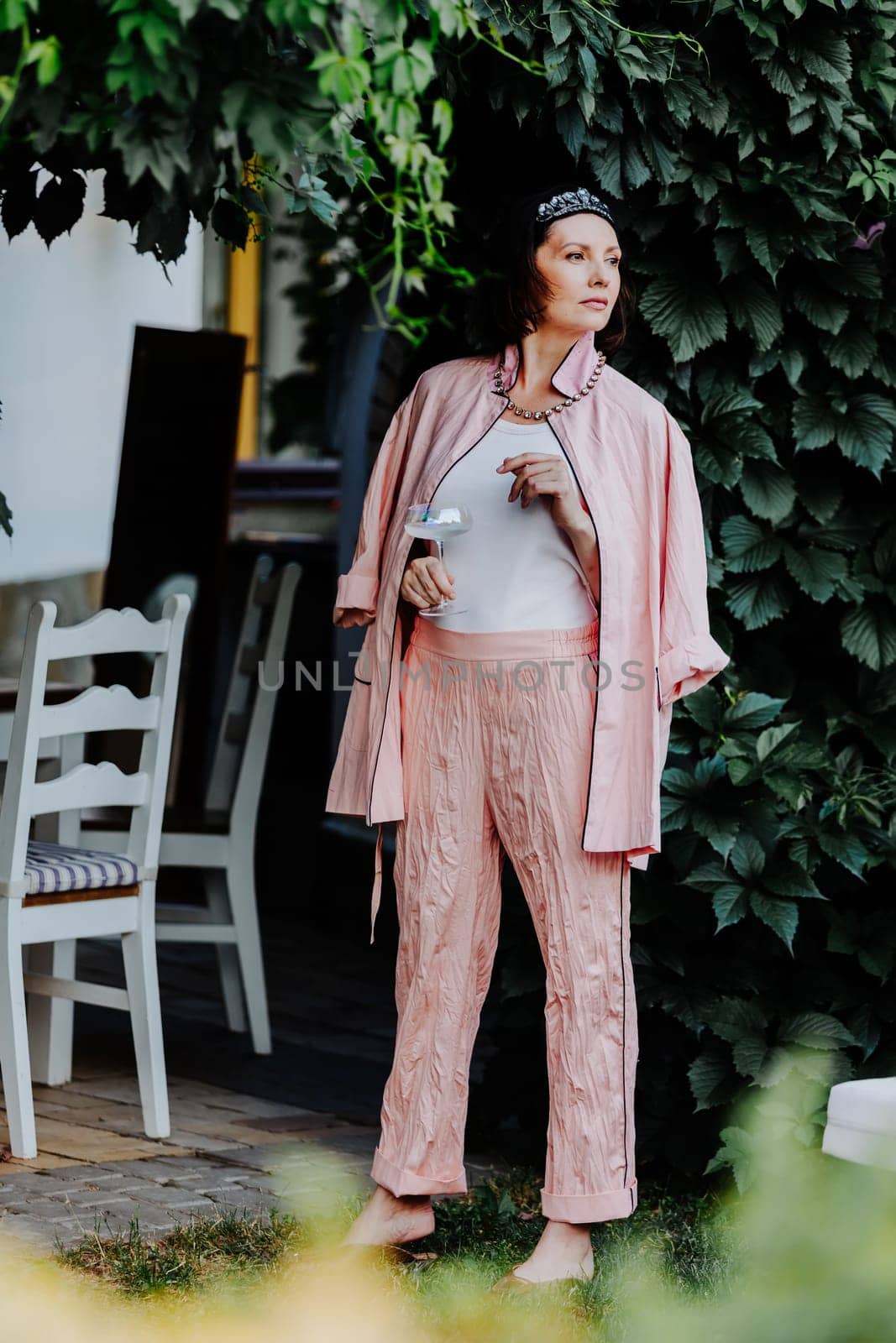Fashionable woman dressed in a stylish light pink ensemble, including a satin jacket and matching pants, is standing outdoors under a canopy of leaves, holding a glass
