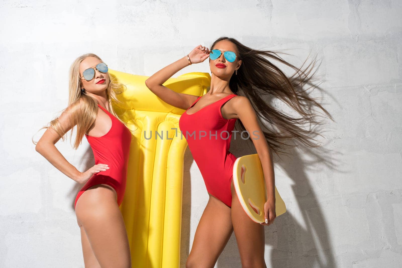 Two sexy girls wearing sunglasses, red swimsuit and summer hat posing in studio near brick white wall holding yellow swimming air mattress