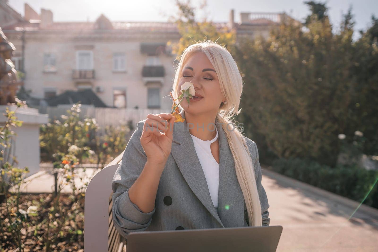 A blonde woman is sitting on a chair and eating a piece of broccoli. She is also using a laptop