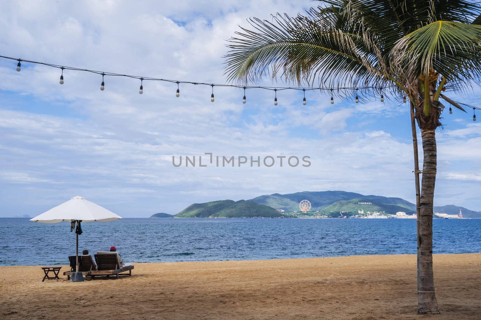 A sandy beach with a palm tree and tourists relaxing under beach umbrellas by the sea in Nha Trang resort in Vietnam overlooking Vinpearl Island