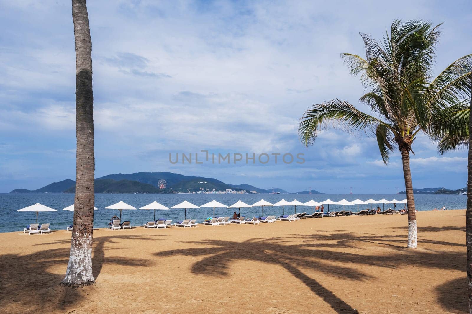 sandy beach with palm trees and tourists relaxing under beach umbrellas by the sea in Nha Trang resort in Vietnam overlooking Vinpearl Island