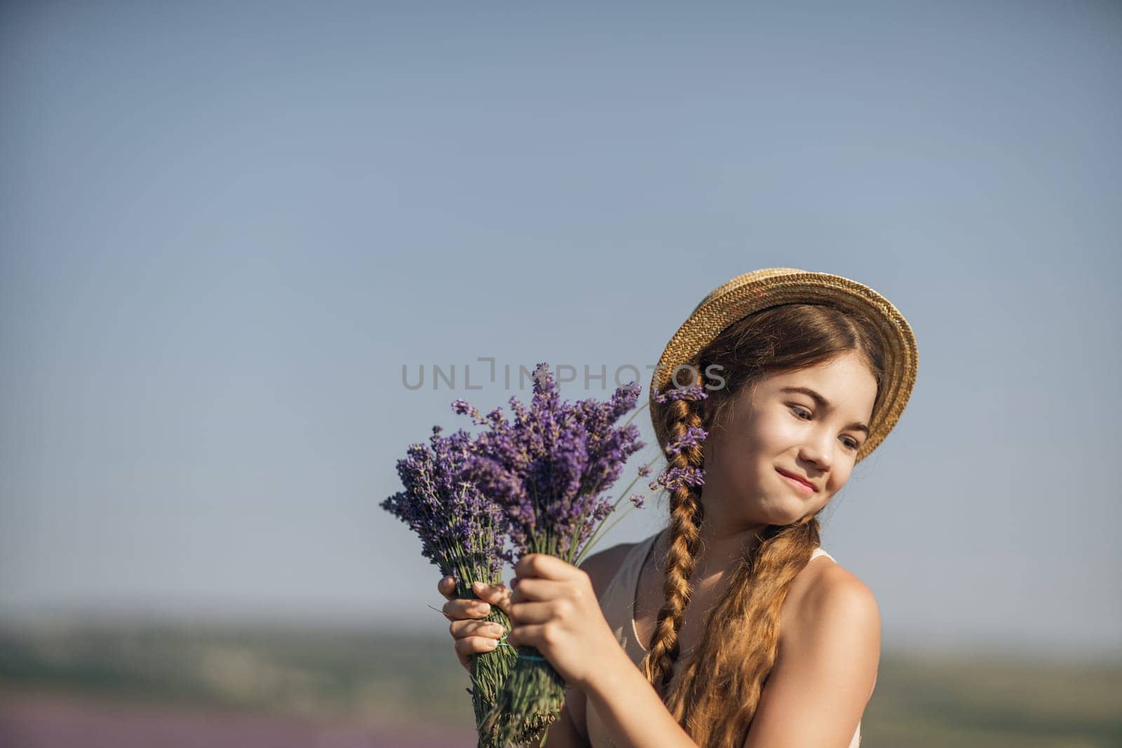 girl is holding a bunch of lavender purple flowers in her hands and wearing a straw hat. She is smiling and she is enjoying herself. The scene is set in a field of lavender, which adds to the peaceful