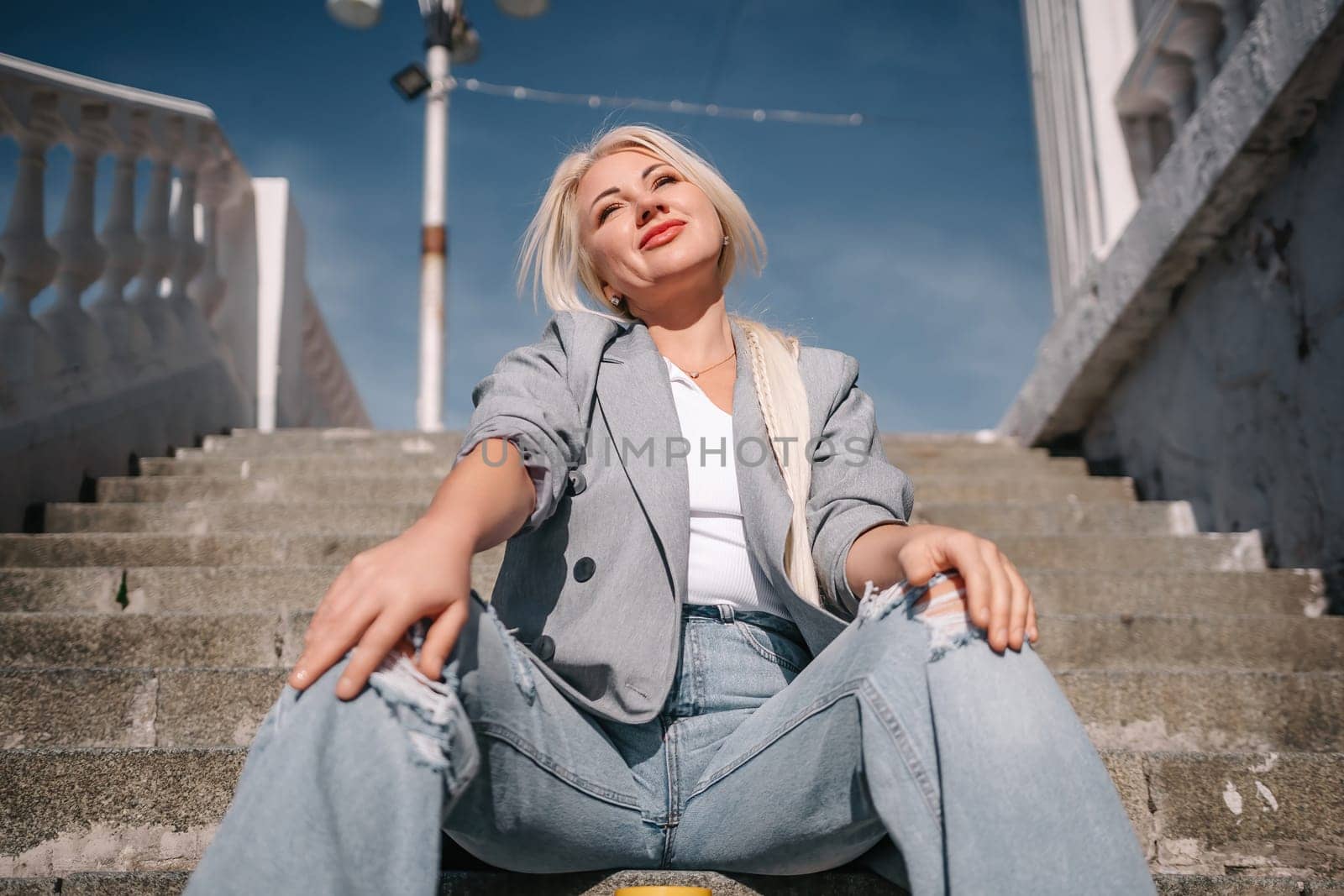 A woman in a gray jacket and blue jeans sits on a set of stairs. She is smiling and she is enjoying herself