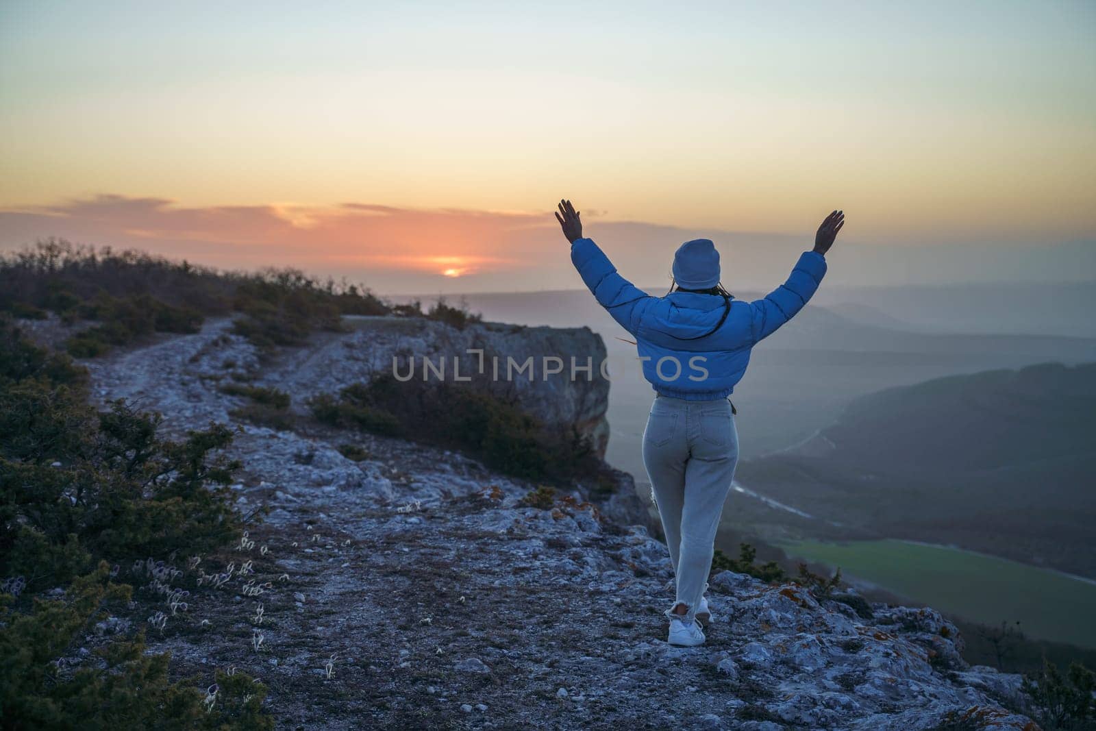 Woman hiker open arms on top of sunrise mountain. The girl salutes the sun, wearing a blue jacket, white hat and white jeans. Conceptual design