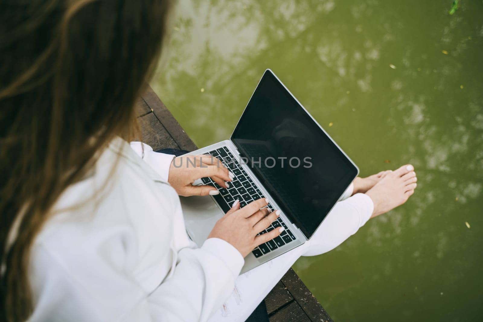 A woman is sitting on a dock with a laptop open in front of her. She is typing on the keyboard and she is focused on her work. Concept of productivity and concentration