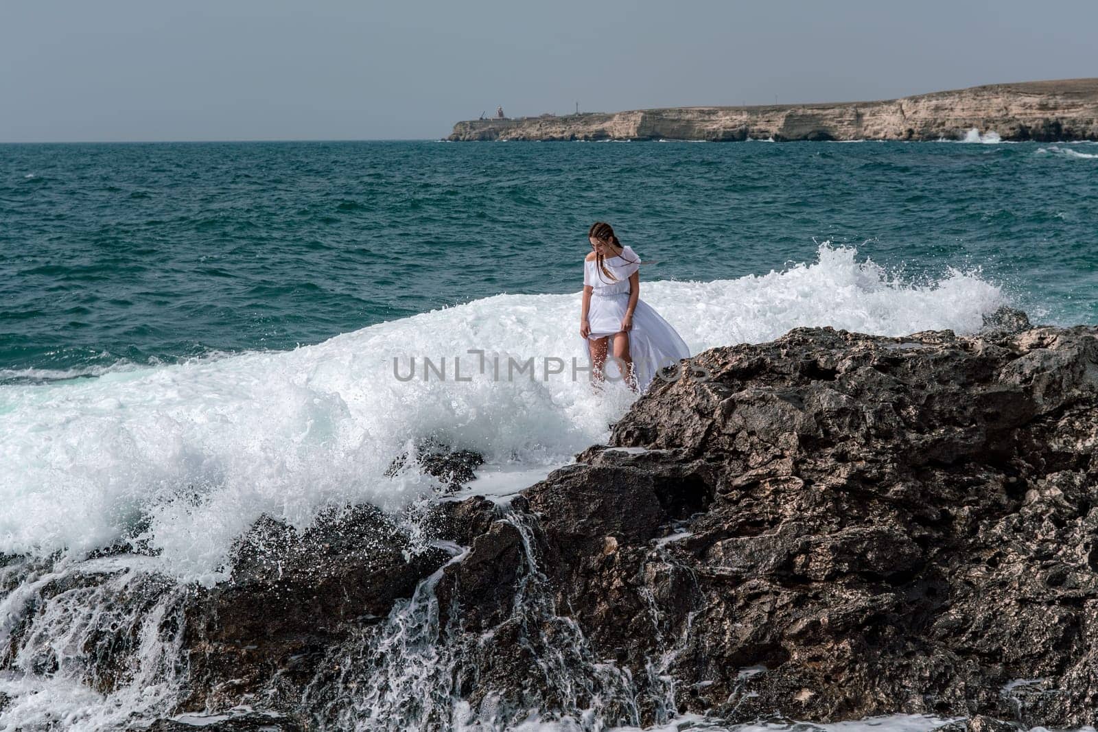 A woman stands on a rock in the sea during a storm. Dressed in a white long dress, the waves break on the rocks and white spray rises