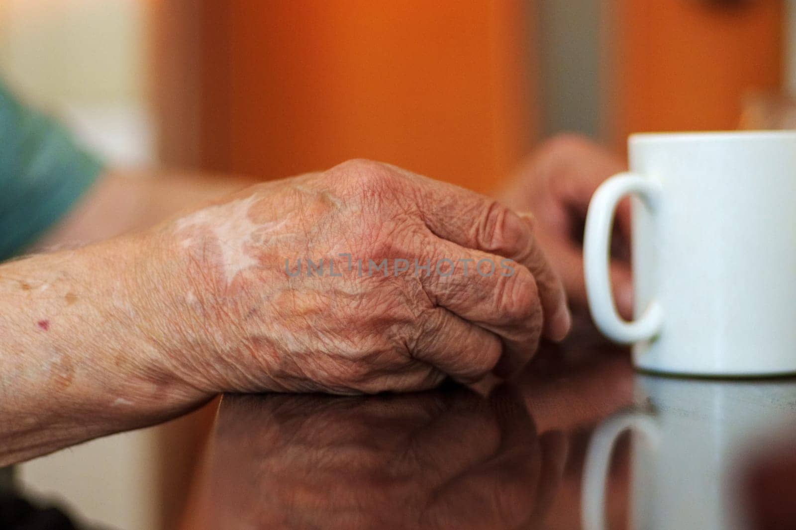 An elderly person hands are gently resting on a table near a white mug.