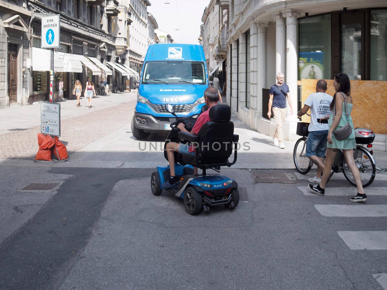 Cremona, Italy August 31st 2024 Senior man driving a mobility scooter crossing the street in the city center with pedestrians and a van in the background