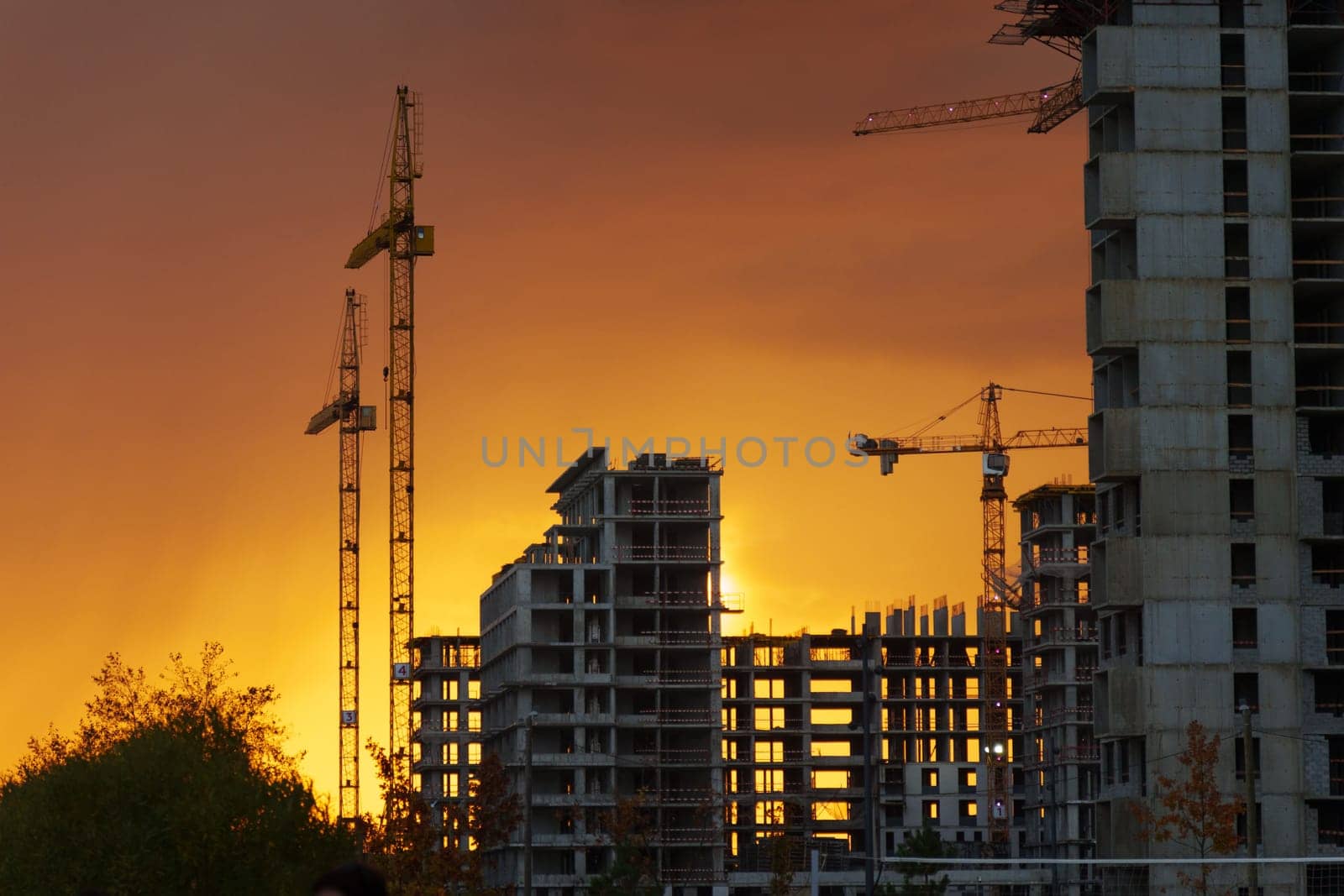 As the sun sets, a striking orange glow envelops the skyline, highlighting cranes and unfinished structures, showcasing the energy of urban development.