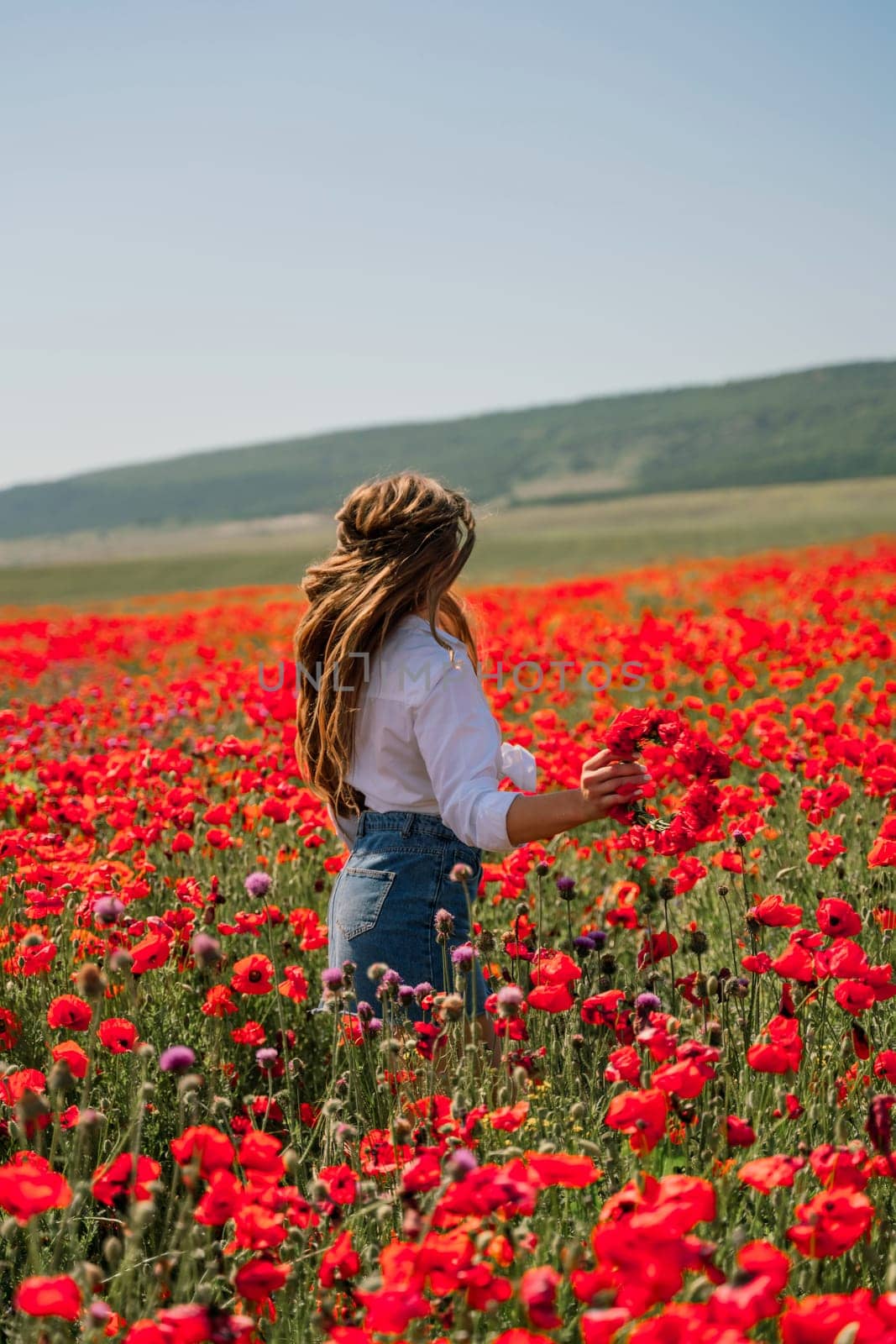 Happy woman in a poppy field in a white shirt and denim skirt with a wreath of poppies on her head posing and enjoying the poppy field