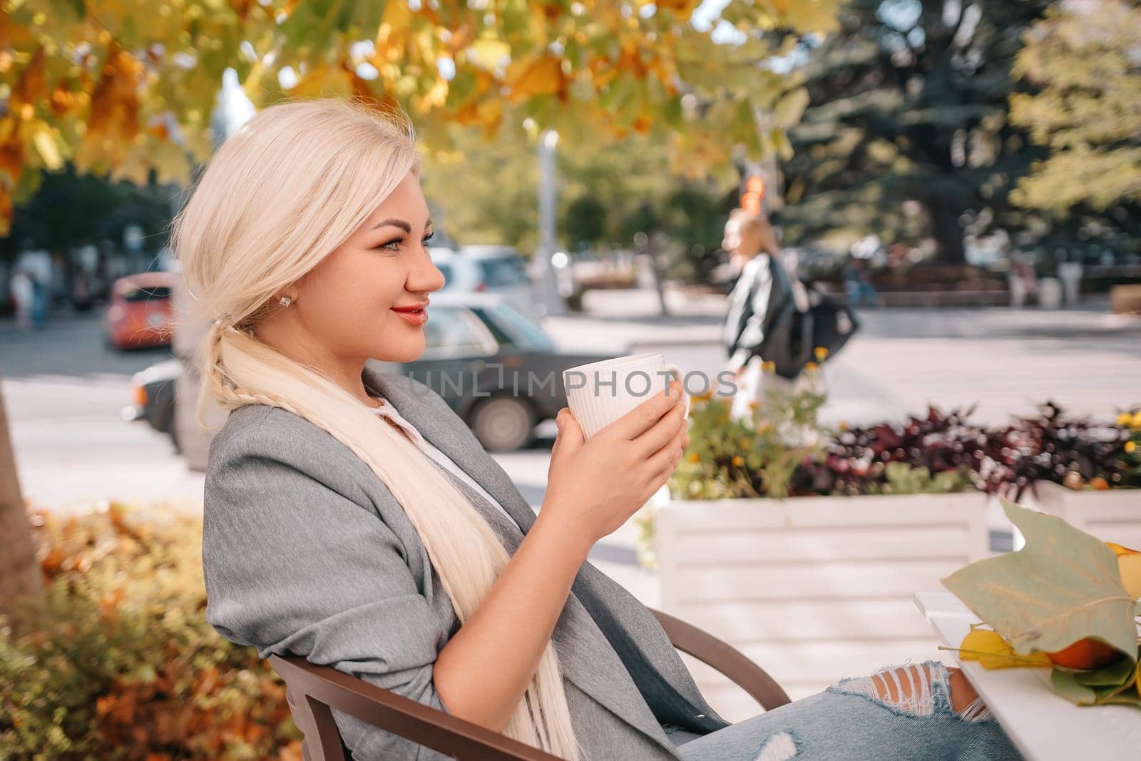 A blonde woman is drinking coffee while sitting under a tree. The scene is peaceful and relaxing, with the woman enjoying her coffee and the natural surroundings