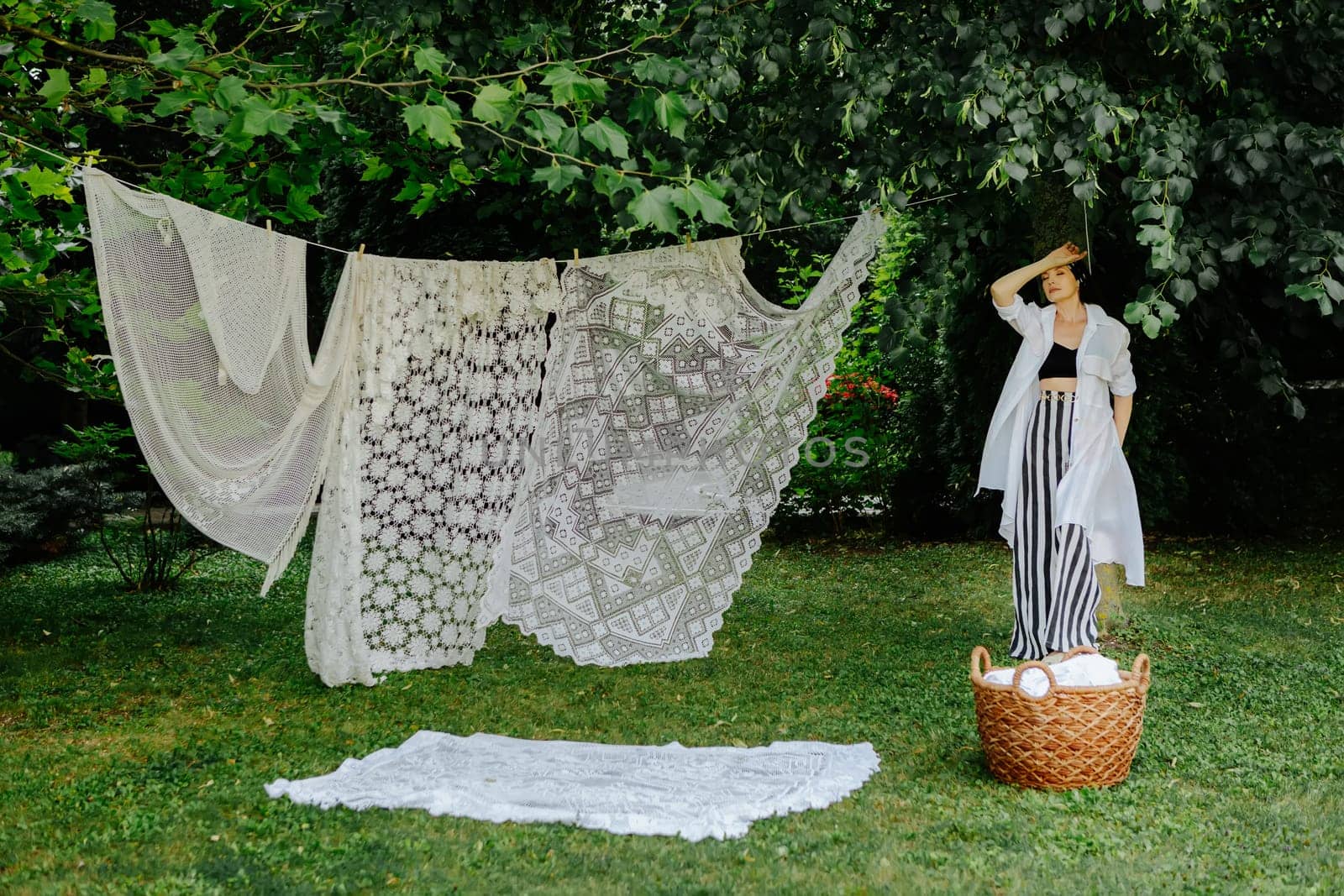 Woman stands in a lush green garden, hanging intricate lace fabric on a clothesline.