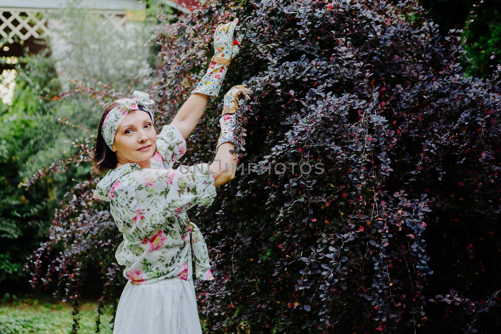 Woman dressed in a floral gardening outfit is pruning a large shrub in a lush garden.