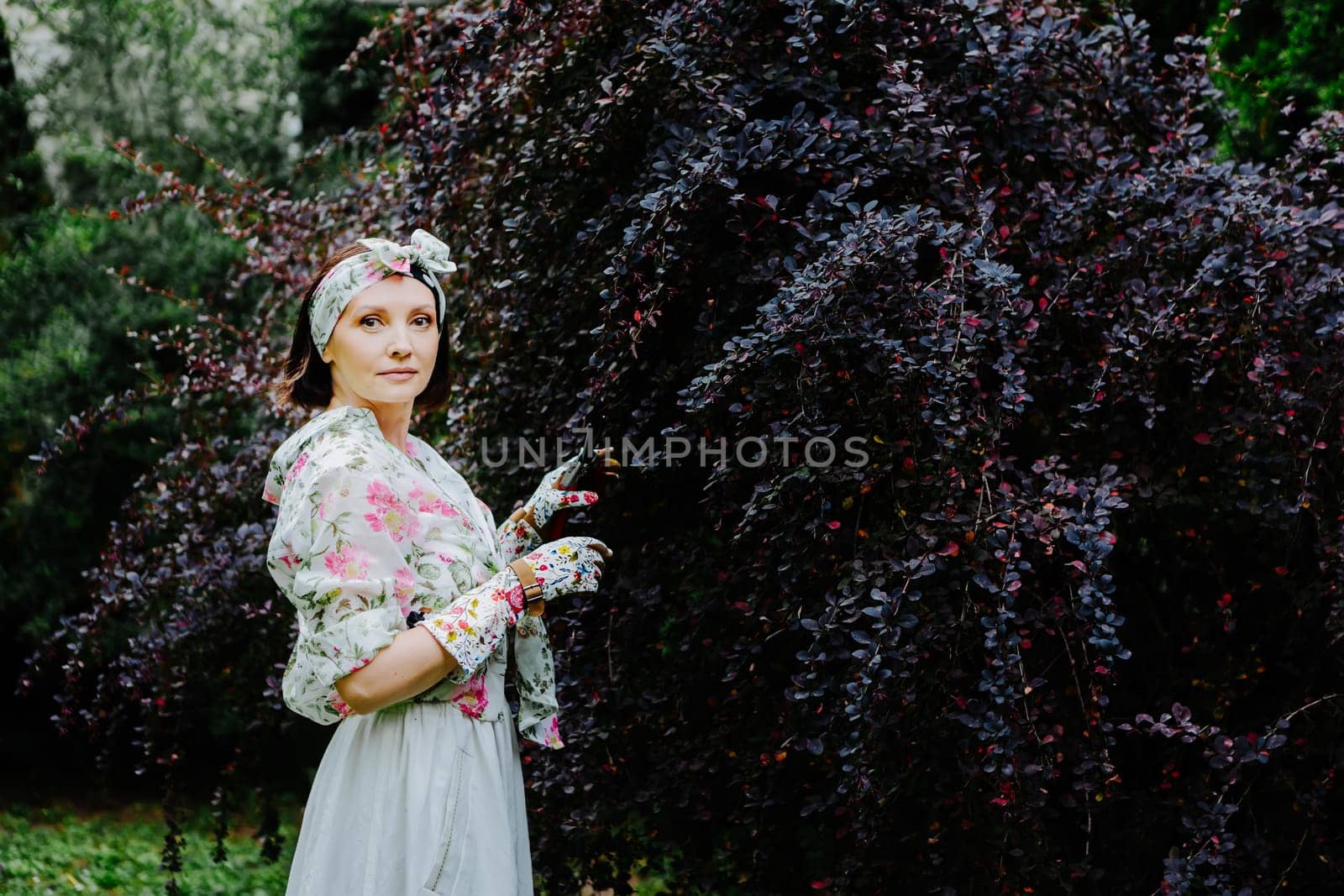 Woman dressed in a floral gardening outfit is pruning a large shrub in a lush garden.
