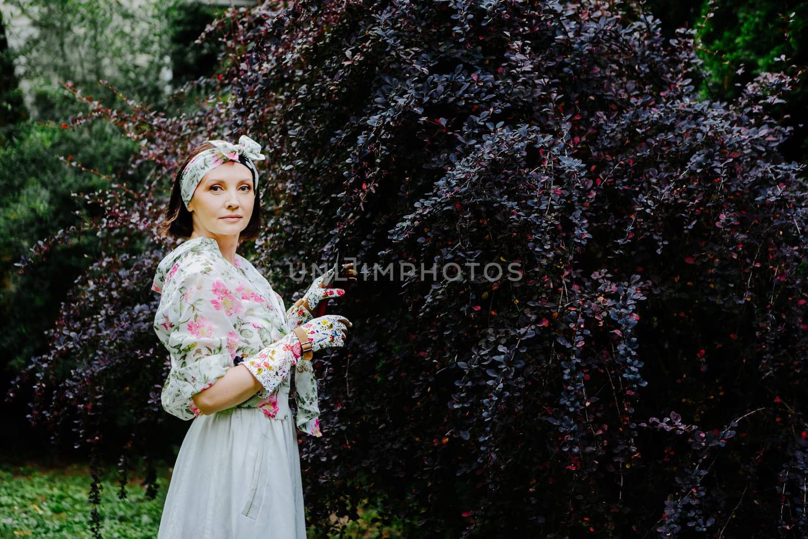 Woman dressed in a floral gardening outfit is pruning a large shrub in a lush garden.