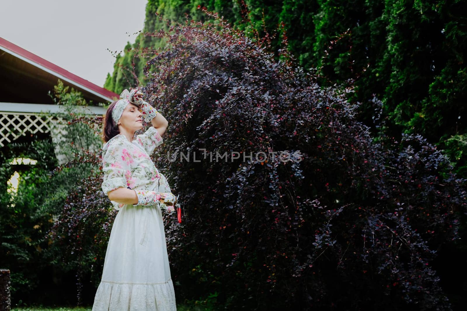 Woman dressed in a floral gardening outfit is pruning a large shrub in a lush garden.