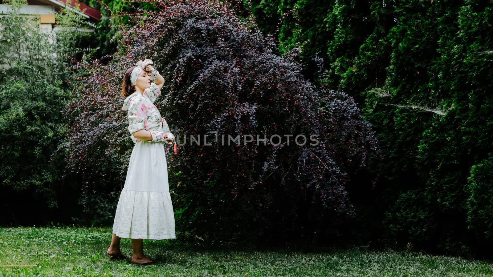 Woman dressed in a floral gardening outfit is pruning a large shrub in a lush garden.
