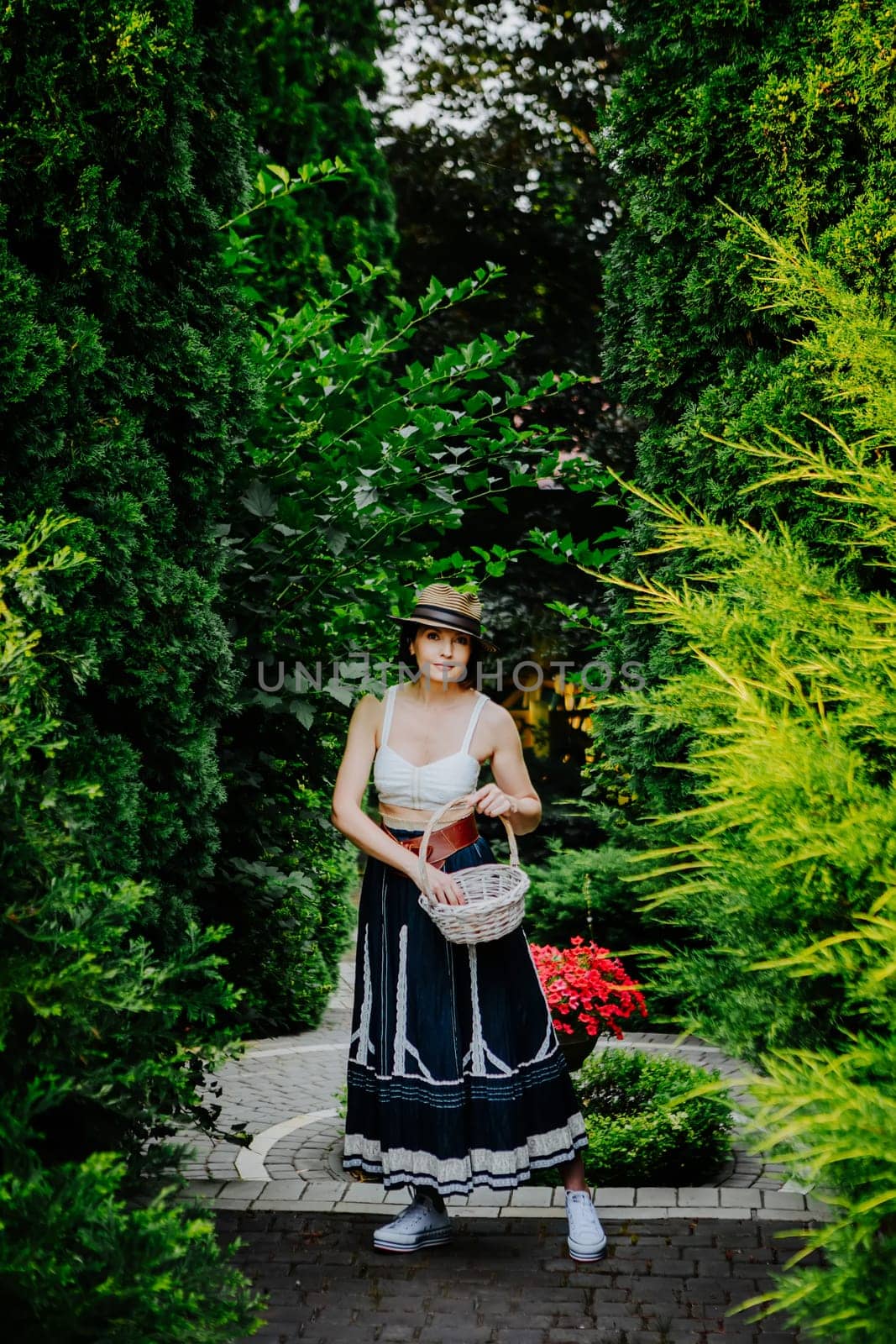 A woman in a stylish summer outfit and a striped sun hat is collecting eggs in a wicker basket in a lush garden.