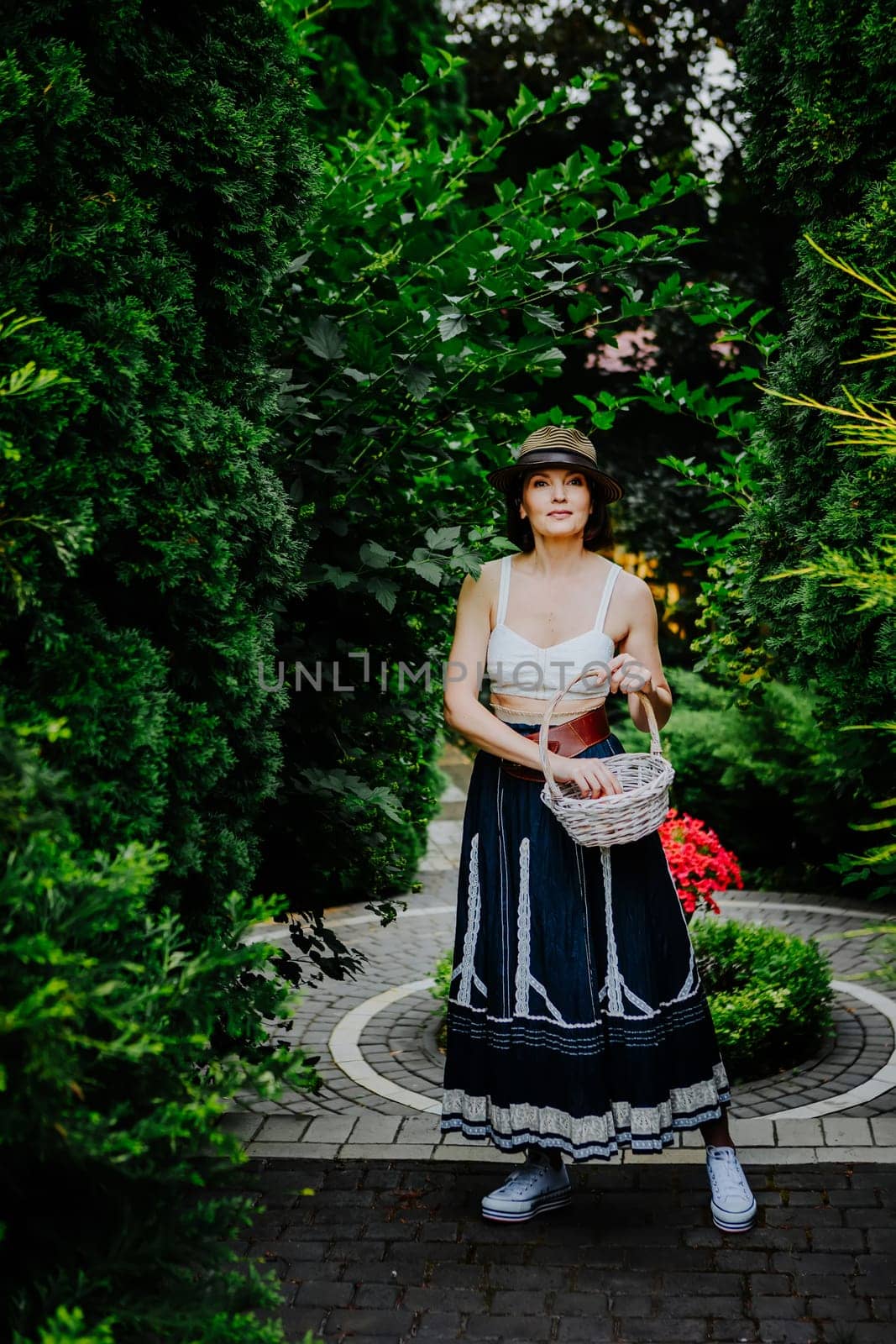 A woman in a stylish summer outfit and a striped sun hat is collecting eggs in a wicker basket in a lush garden.