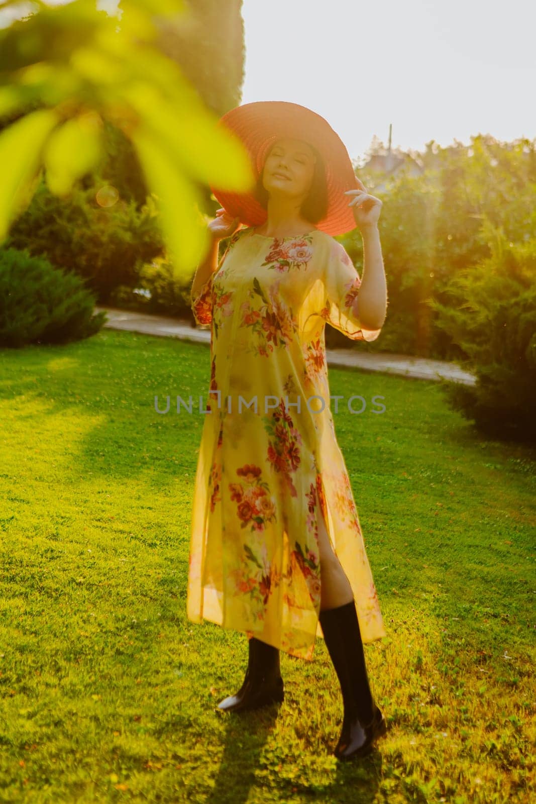 A woman wearing a floral dress and a large pink sun hat stands in a lush garden during golden hour. She holds the brim of her hat, casting a soft shadow over her face as the sunlight illuminates the scene.