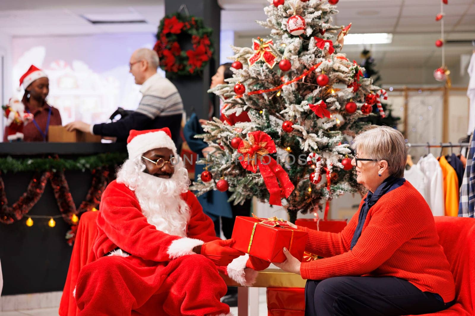 Older client sitting with santa claus in shop, receiving amazing christmas gift from employee next to festive ornaments. African american worker spreading holiday cheer in shopping mall.
