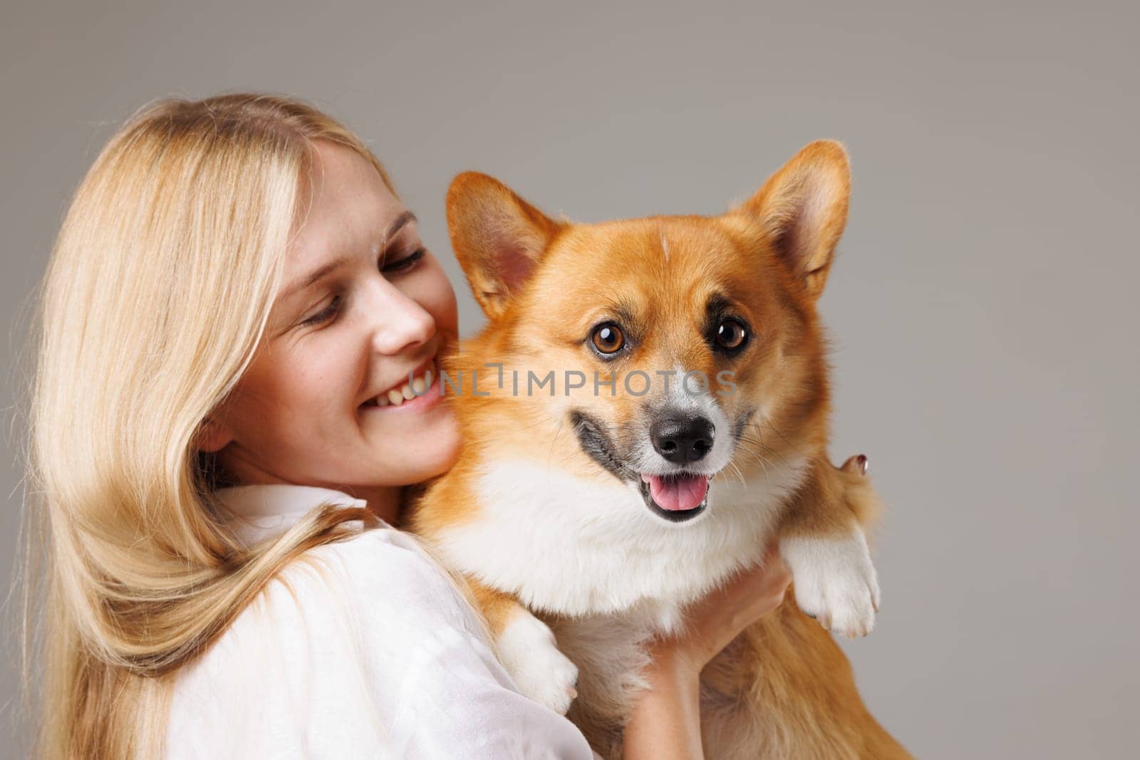 A woman with long blonde hair smiles while holding a happy corgi dog in her arms. The background is neutral, emphasizing the joyful interaction between the woman and her pet.