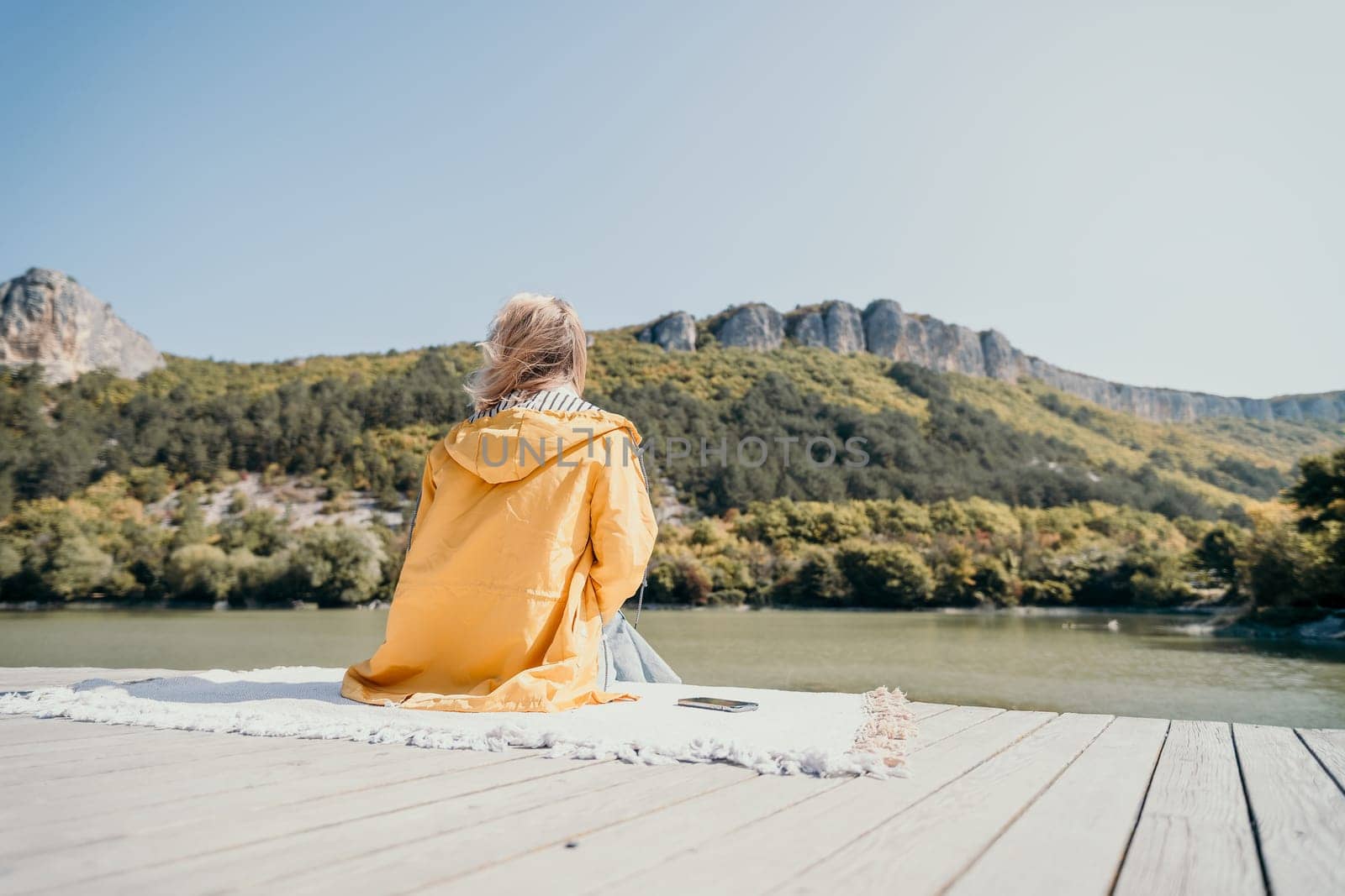 A woman in a yellow jacket sits on a wooden dock, gazing towards a mountain range by a calm lake.