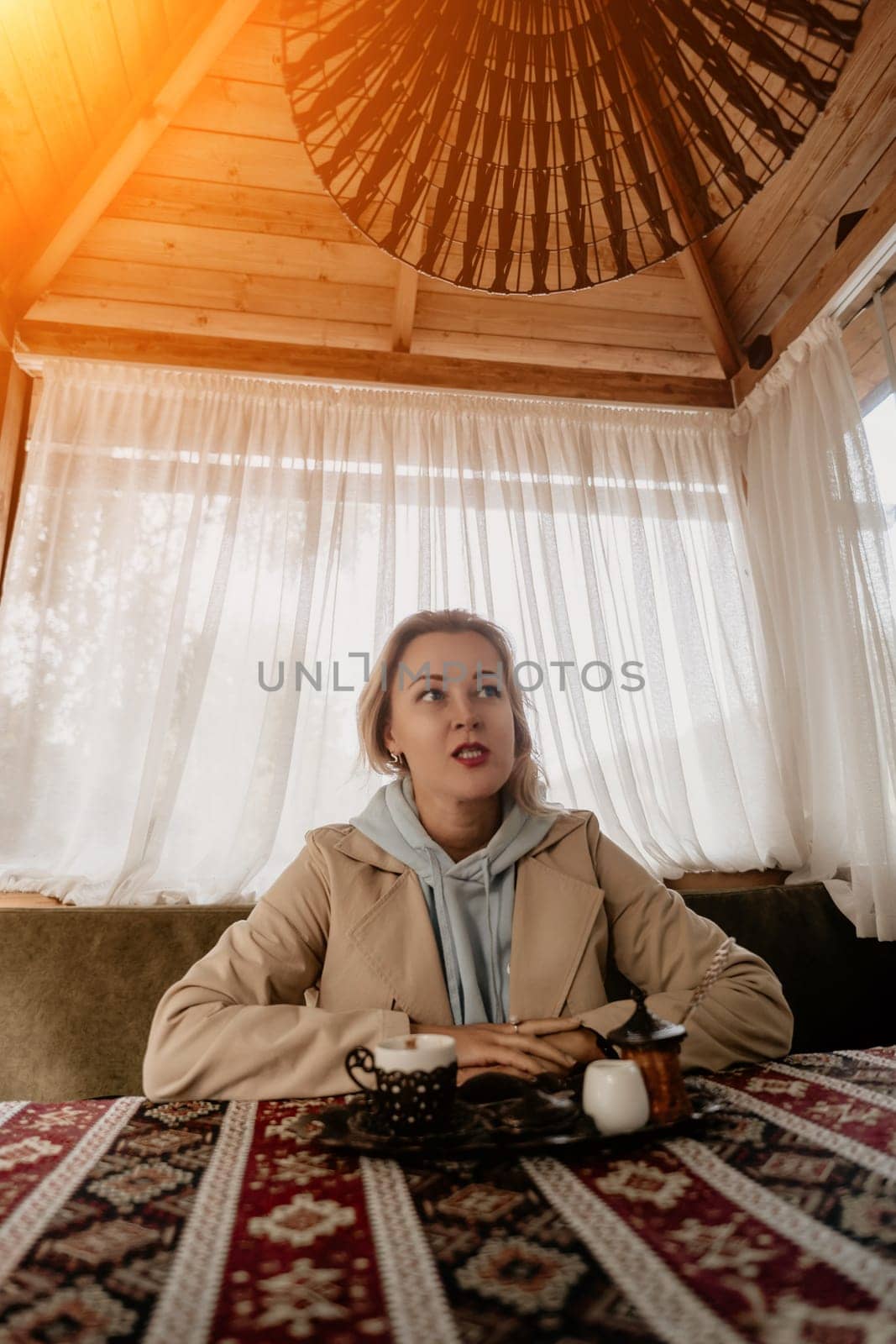 A young woman in a light blue hoodie and beige coat sits at a table in a cozy cafe, looking thoughtfully towards the left, a cup of coffee in front of her.