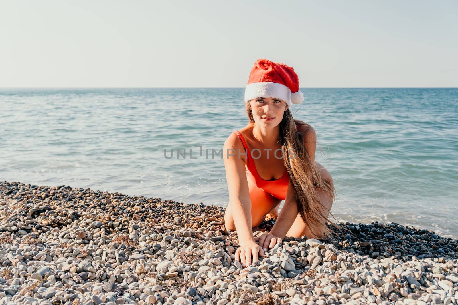 A young woman in a red swimsuit and a Santa hat poses on a pebble beach in front of the ocean.