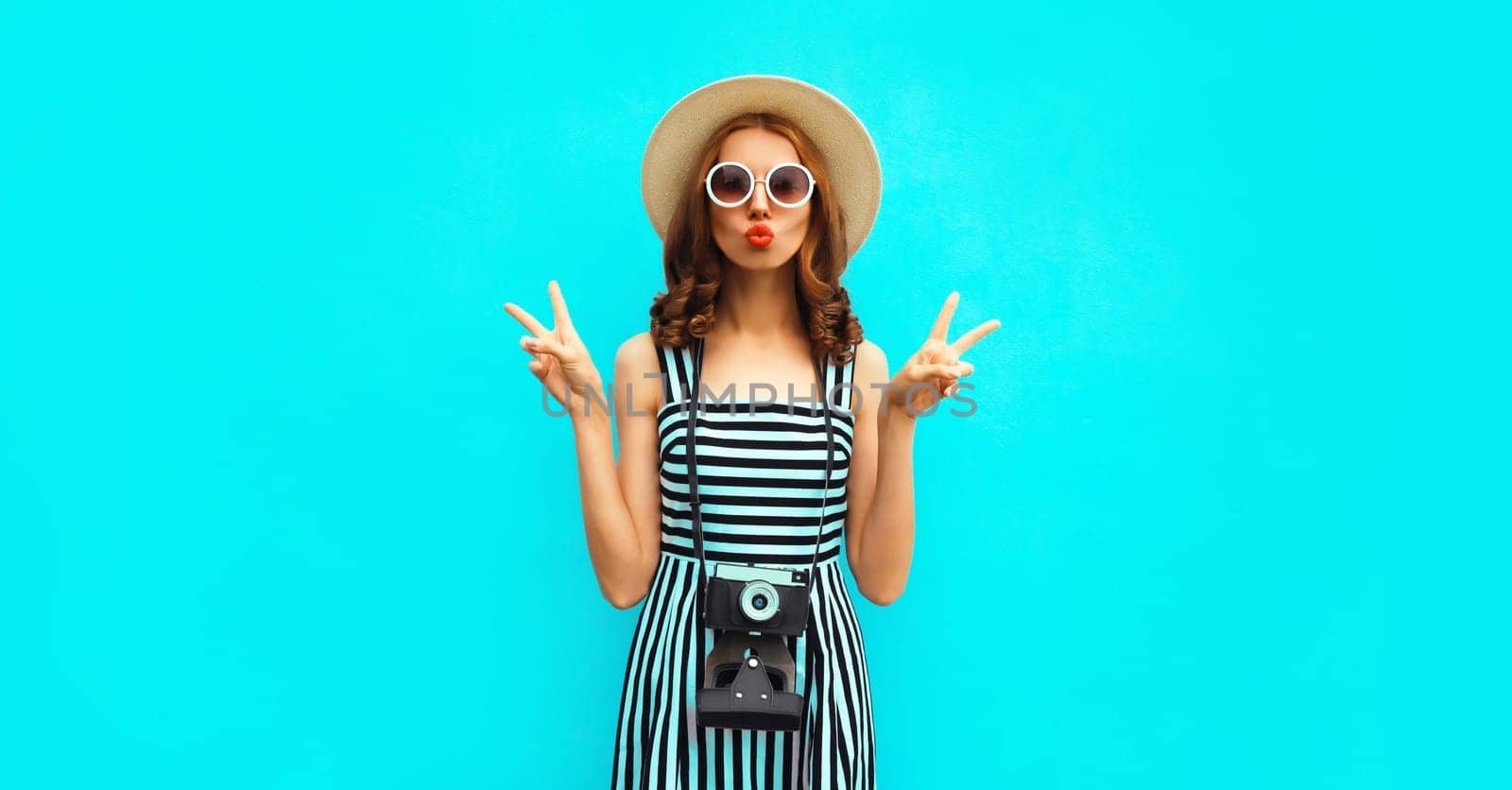 Summer portrait of happy smiling young woman photographer with vintage film camera wearing straw hat, dress, sunglasses on blue background