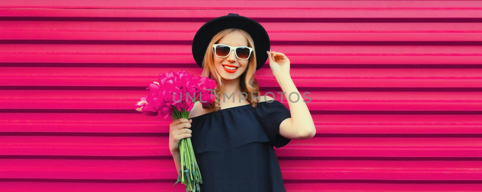 Sweet portrait of beautiful happy smiling woman with bouquet of pink rose flowers posing in black round hat on colorful background
