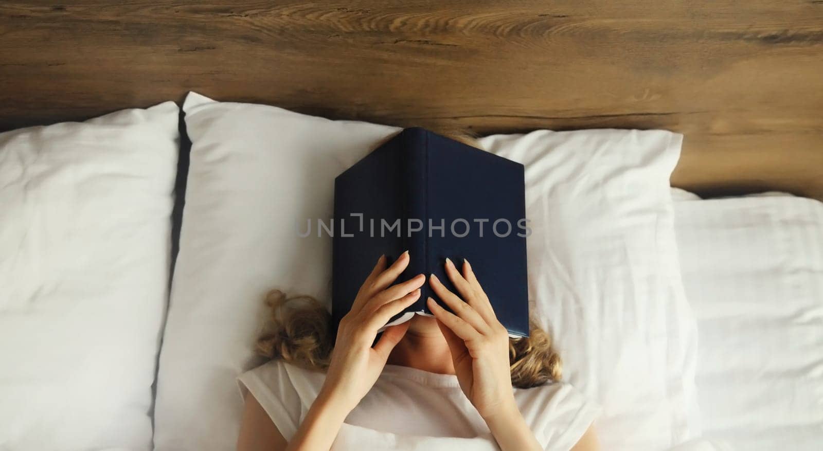 Young sad tired woman student covering her face with open book with blank cover while lying on the bed at home